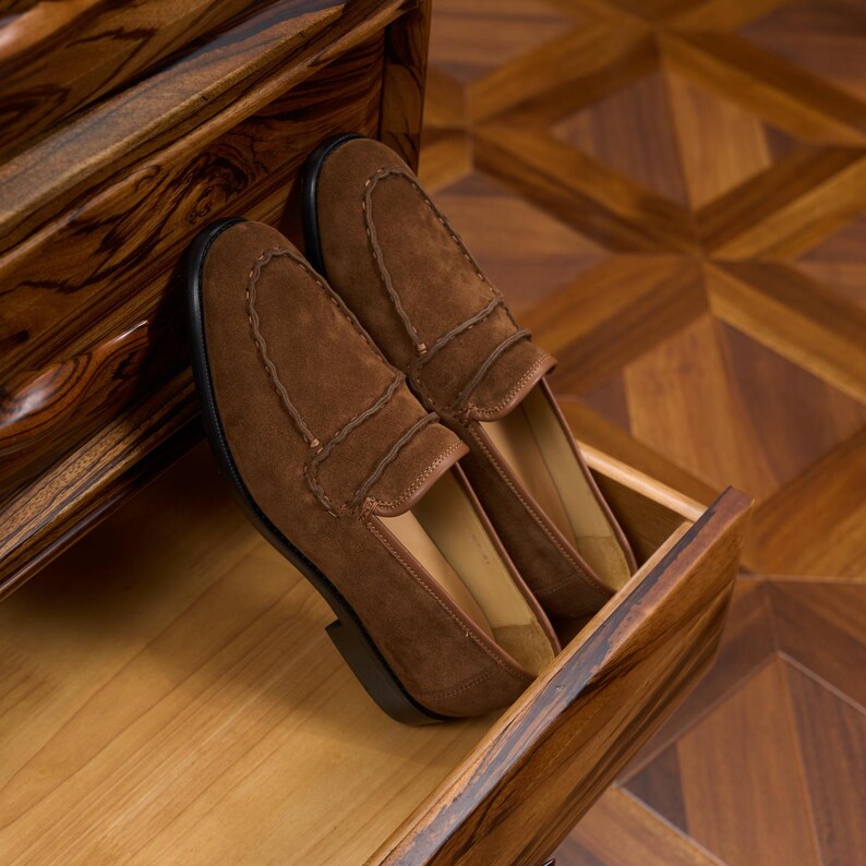 May include: A pair of brown suede loafers featuring a moccasin-style stitched toe seam and a low heel. The shoes have a smooth tan interior lining and a dark brown sole, presented with a clean finish and visible texture on the suede material.