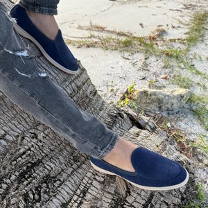 May include: Navy blue suede slip-on loafers with a cream-colored sole. The shoes are worn with gray denim jeans. The loafers have a classic design, suitable for casual wear. The image is taken outdoors, with a natural background.