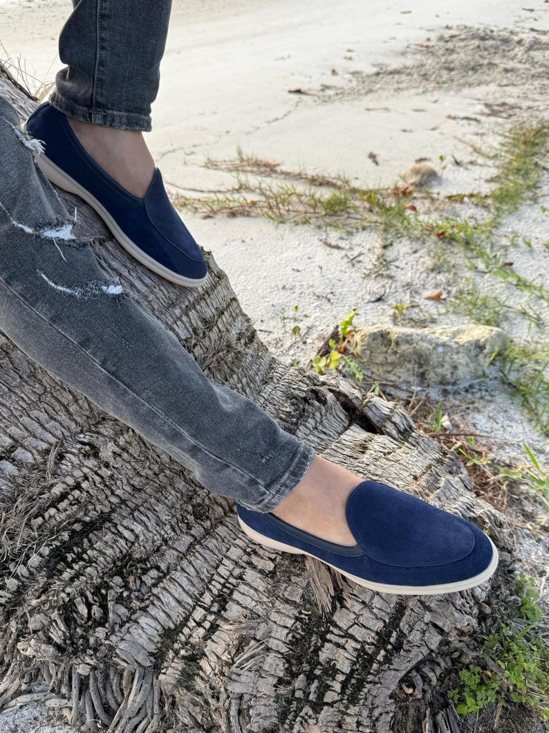 May include: Navy blue suede slip-on loafers with a cream-colored sole. The shoes are worn with gray denim jeans. The loafers have a classic design, suitable for casual wear. The image is taken outdoors, with a natural background.