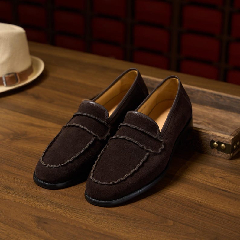 May include: A pair of dark brown suede loafers featuring a scalloped edge detail along the vamp strap and the moccasin-style toe seam. The shoes have a smooth tan interior lining and a solid black sole, presented as a matching set of footwear.