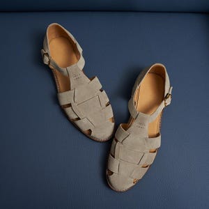 May include: A pair of gray suede sandals with a woven design. The sandals have a buckle closure and a light brown insole. The shoes are displayed on a blue surface.