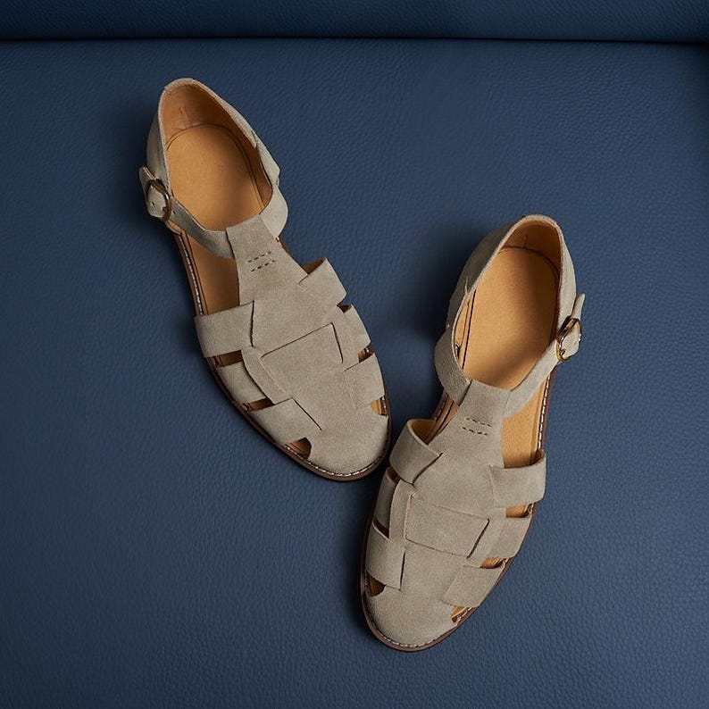 May include: A pair of gray suede sandals with a woven design. The sandals have a buckle closure and a light brown insole. The shoes are displayed on a blue surface.