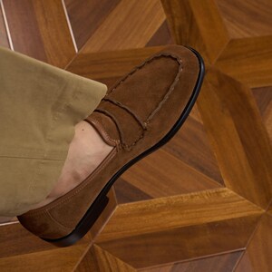 May include: A brown suede loafer with a moccasin-style stitched toe seam and a horizontal strap across the vamp. The shoe features a low heel and a dark sole, worn with tan trousers. The design includes visible stitching along the upper edge.