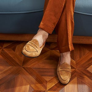May include: A pair of tan suede penny loafers featuring a classic strap across the vamp, a rounded toe, and a low-profile silhouette. The shoes are worn with rust-colored trousers that have a cuffed hem, showcasing the soft texture of the suede.