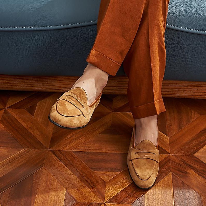 May include: A pair of tan suede penny loafers featuring a classic strap across the vamp, a rounded toe, and a low-profile silhouette. The shoes are worn with rust-colored trousers that have a cuffed hem, showcasing the soft texture of the suede.