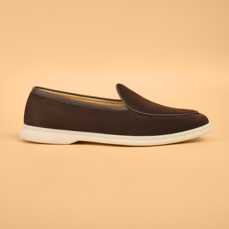 May include: A brown suede loafer with a rounded toe and a white sole. The shoe has a dark brown trim around the opening and a low heel. The shoe is designed for comfort and style.