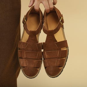 May include: A pair of brown leather sandals with a woven design. The sandals have a T-strap and a buckle closure. The soles are black with a light brown interior.