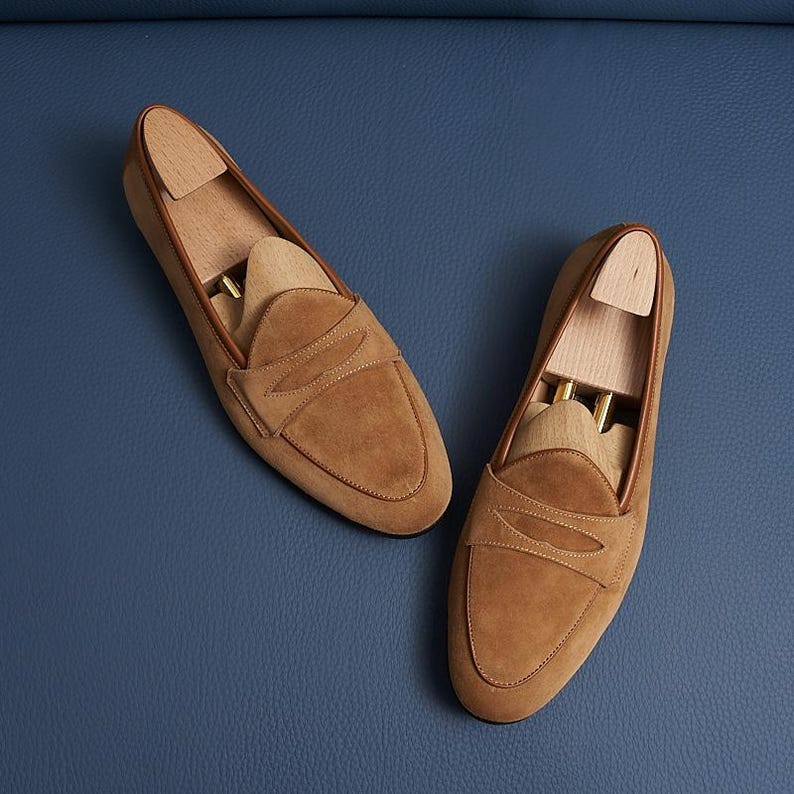 May include: A pair of tan suede penny loafers featuring a classic moccasin-style construction with a horizontal strap across the vamp. Each shoe contains a light-colored wooden shoe tree insert with a metal spring mechanism for shape retention.