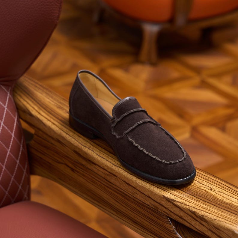 May include: A dark brown suede loafer featuring a rounded toe, a classic penny strap across the vamp, and a scalloped edge detail along the stitching. The shoe has a smooth interior lining and a dark, low-profile sole, showcasing a textured finish.