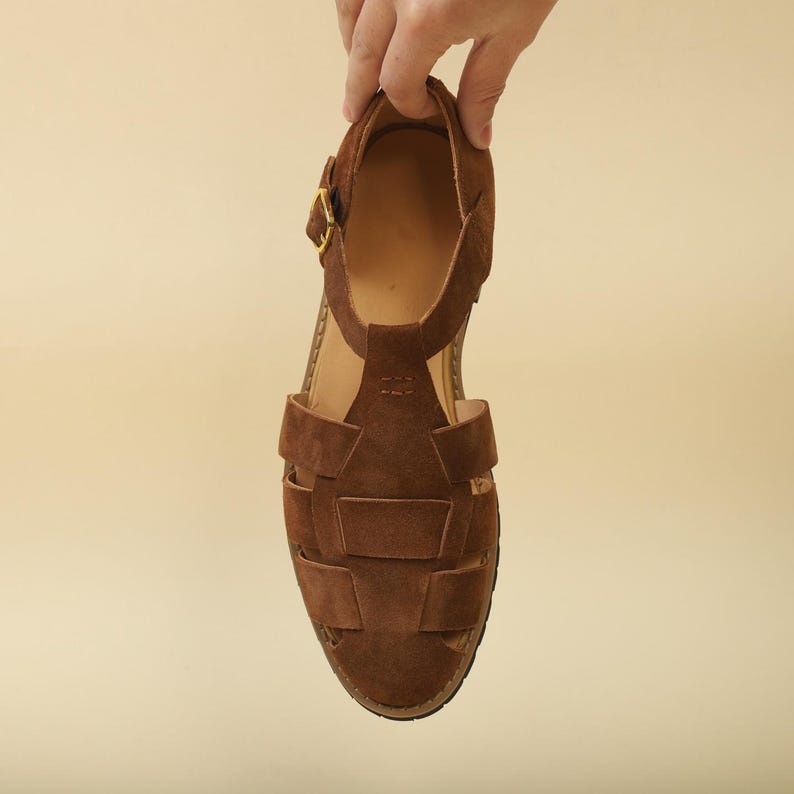 May include: A brown suede sandal with a woven design. The sandal features a T-strap and a gold-colored buckle. The sole is a dark brown color. The sandal is being held up by a hand.