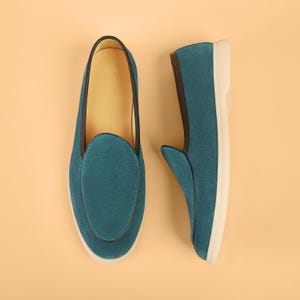 May include: Teal suede slip-on shoes with a rounded toe and a white sole. The shoes have a black trim and a leather interior. The shoes are displayed on a light orange background.
