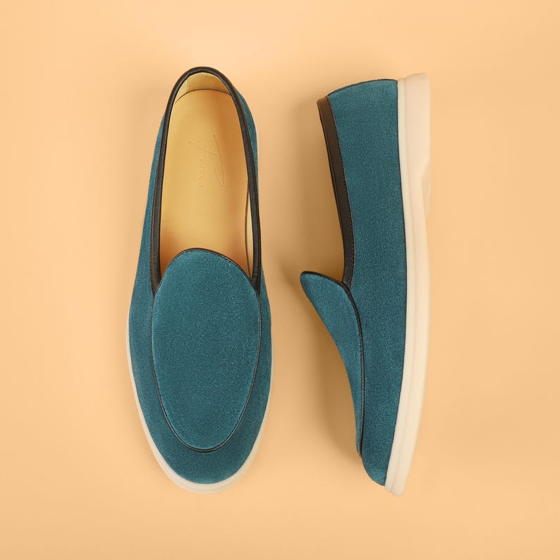 May include: Teal suede slip-on shoes with a rounded toe and a white sole. The shoes have a black trim and a leather interior. The shoes are displayed on a light orange background.
