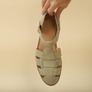 May include: A light gray sandal with a woven design on the front. The sandal has a strap around the ankle and a gold-colored buckle. The sole is brown, and the sandal is held up by a hand.