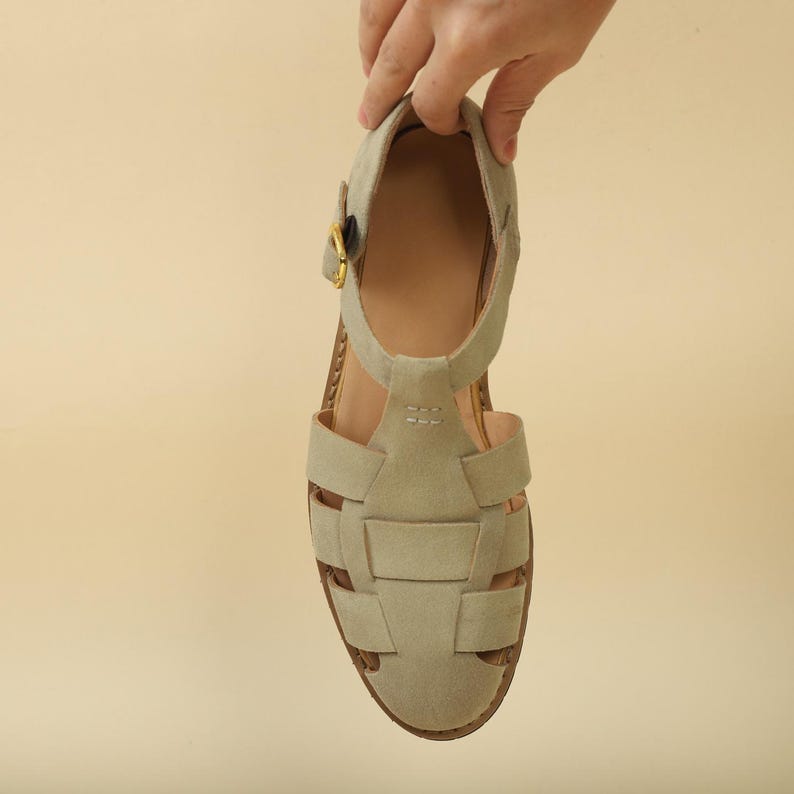 May include: A light gray sandal with a woven design on the front. The sandal has a strap around the ankle and a gold-colored buckle. The sole is brown, and the sandal is held up by a hand.