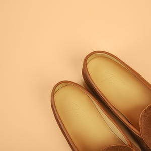 May include: A pair of brown suede loafers with leather trim and a light tan interior. The shoes have a classic design and are set against a peach-colored background. The brand name is subtly embossed on the insole.