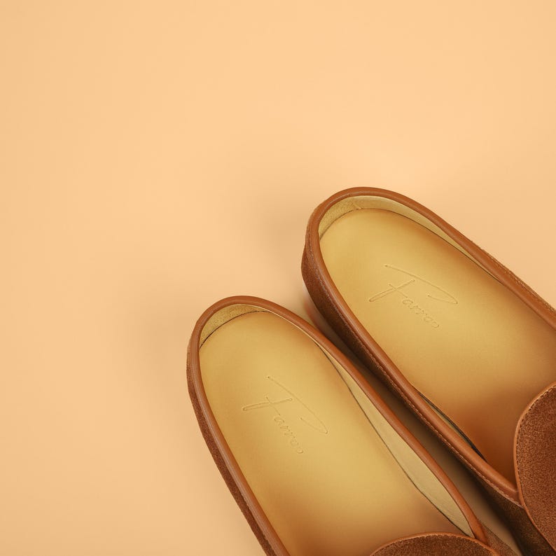 May include: A pair of brown suede loafers with leather trim and a light tan interior. The shoes have a classic design and are set against a peach-colored background. The brand name is subtly embossed on the insole.