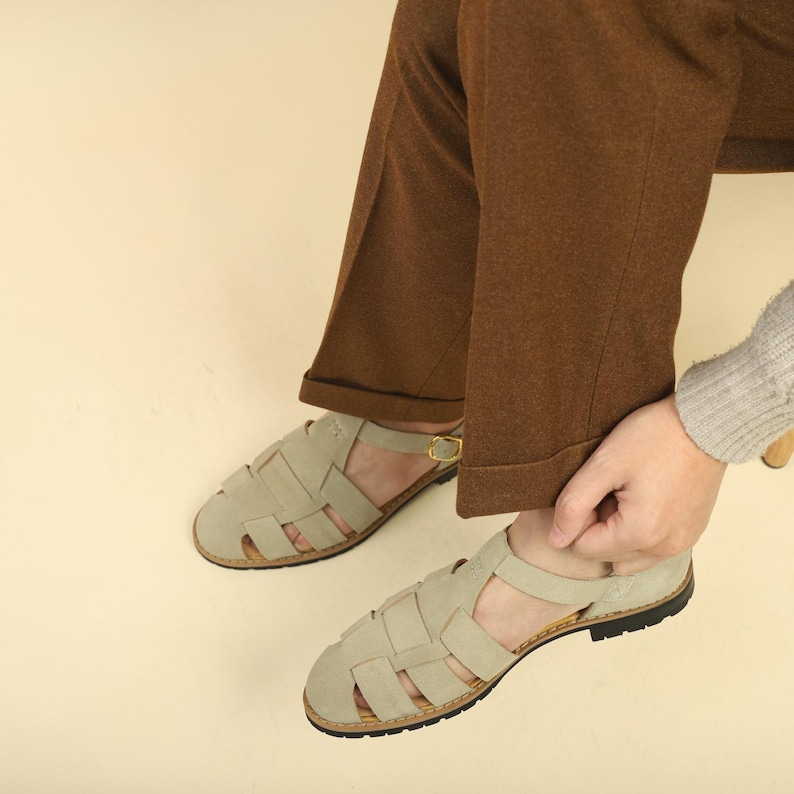 May include: A pair of light beige suede sandals with a woven design. The sandals have a strap around the ankle with a gold-colored buckle and a cork sole. The sandals are worn with brown pants.