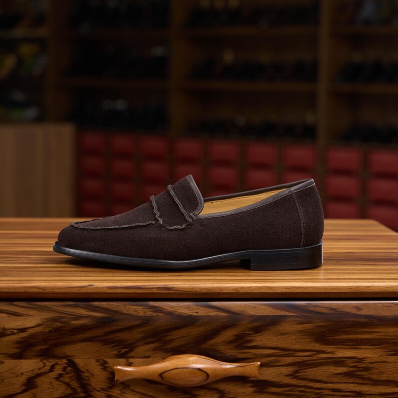 May include: A dark brown suede loafer featuring a classic penny strap across the vamp, a rounded toe, and a low black heel. The shoe has a smooth leather trim along the collar and a visible tan interior lining, presented in a side profile view.