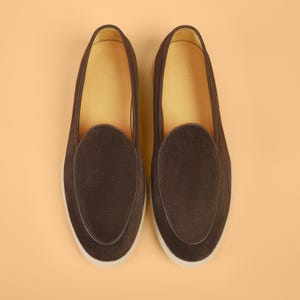 Op de afbeelding: Een paar donkerbruine su&egrave;de loafers met een ronde neus en een witte zool. De binnenkant is lichtbruin. De schoenen worden gepresenteerd op een lichtoranje achtergrond.