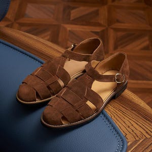 May include: A pair of brown suede sandals with a woven design. The sandals have a strap with a gold buckle and are set on a blue surface. The shoes are a size 9.