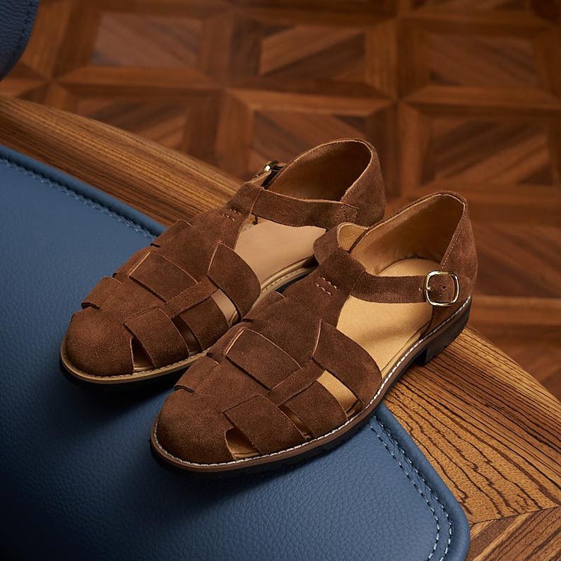 May include: A pair of brown suede sandals with a woven design. The sandals have a strap with a gold buckle and are set on a blue surface. The shoes are a size 9.