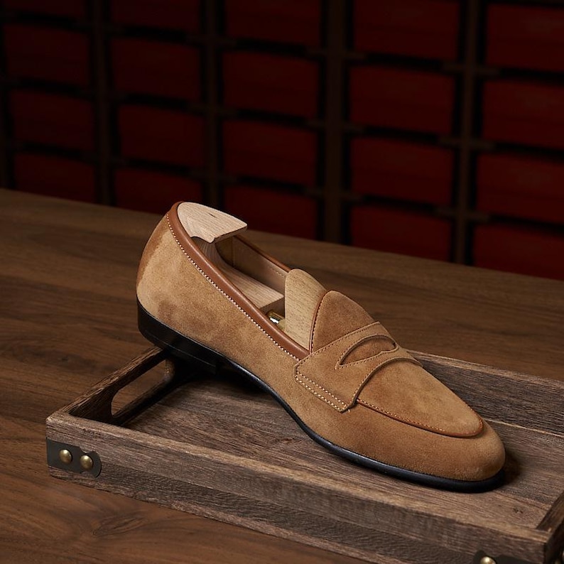 May include: A tan suede penny loafer shoe featuring a smooth leather trim along the collar and a wooden shoe tree inserted inside. The shoe has a classic moccasin-style stitched apron toe and a dark sole, presented as a single footwear item.