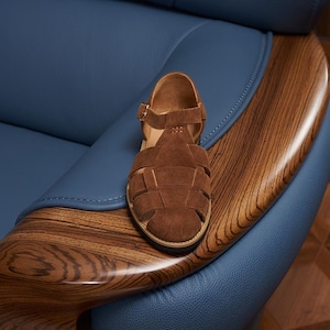 May include: Brown suede sandal with a woven design and a buckle closure. The sandal is placed on a wooden armrest with a blue leather chair in the background. The sandal has a round toe and a flat sole.