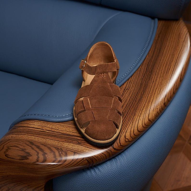 May include: Brown suede sandal with a woven design and a buckle closure. The sandal is placed on a wooden armrest with a blue leather chair in the background. The sandal has a round toe and a flat sole.