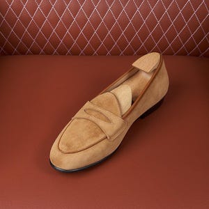 May include: A tan suede penny loafer featuring a rounded toe, a classic saddle strap across the vamp, and a smooth leather trim along the collar. A light-colored wooden shoe tree is inserted inside the shoe to maintain its structured shape.