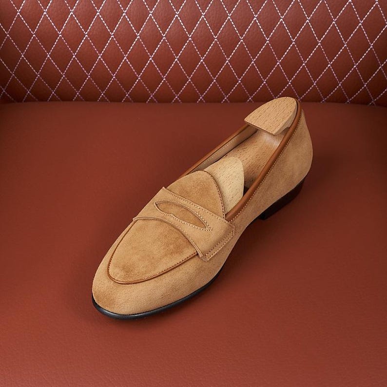 May include: A tan suede penny loafer featuring a rounded toe, a classic saddle strap across the vamp, and a smooth leather trim along the collar. A light-colored wooden shoe tree is inserted inside the shoe to maintain its structured shape.