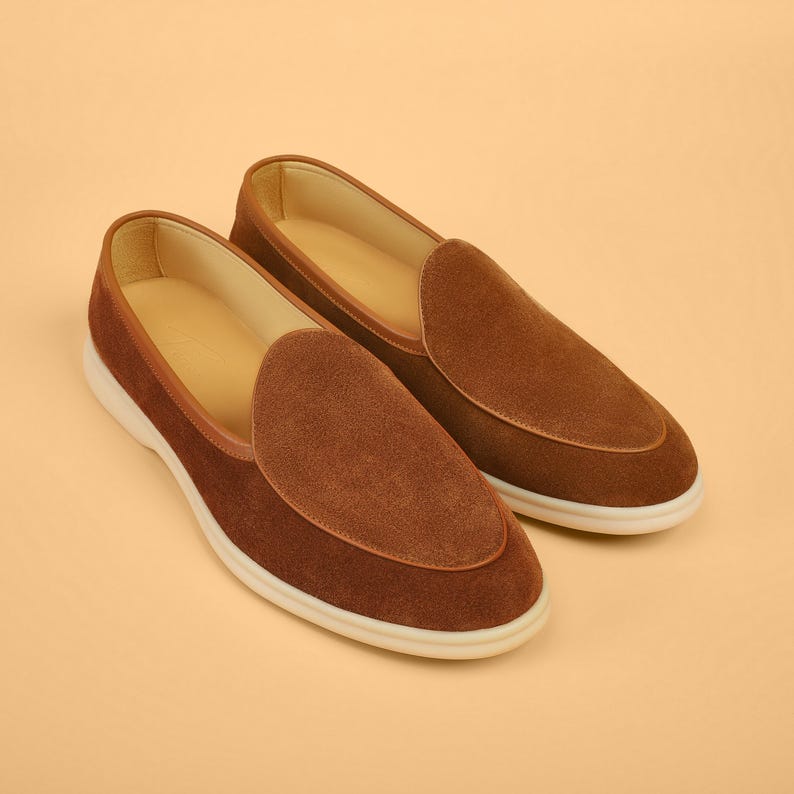May include: A pair of brown suede loafers with a rounded toe and a white sole. The shoes have a classic design with a smooth, soft texture. The interior is a light tan color.