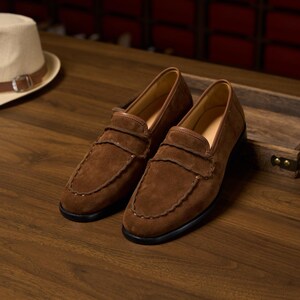 May include: A pair of brown suede loafers featuring a moccasin-style stitched toe seam, a smooth leather collar trim, and a black sole. The shoes have a classic slip-on design with a horizontal strap across the vamp and a rounded toe shape.