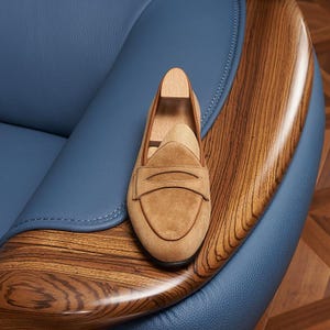 May include: A tan suede penny loafer shoe featuring a rounded toe, a classic saddle strap across the vamp, and a light-colored wooden shoe tree insert. The shoe is constructed with visible stitching along the edges and a smooth, soft texture.