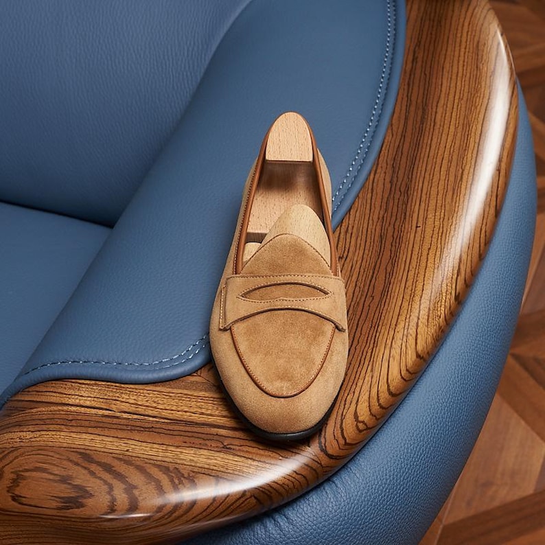 May include: A tan suede penny loafer shoe featuring a rounded toe, a classic saddle strap across the vamp, and a light-colored wooden shoe tree insert. The shoe is constructed with visible stitching along the edges and a smooth, soft texture.