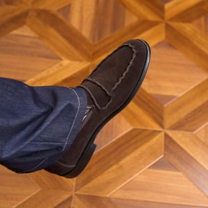 May include: A dark brown suede loafer with a moccasin-style stitched toe seam and a strap across the vamp, worn with dark blue denim jeans. The shoe features a low heel and a rounded toe, showcasing a textured suede finish and visible stitching.