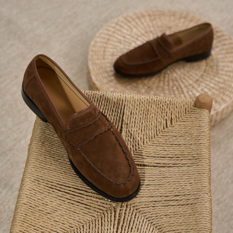 May include: A pair of brown suede loafers featuring a moccasin-style stitched toe seam, a classic penny strap across the vamp, and a low black heel. The shoes have a smooth tan interior lining and a soft, textured exterior finish in a rich brown.