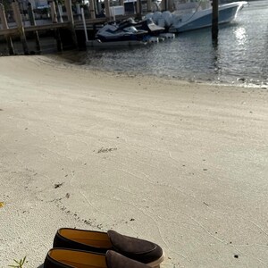 May include: A pair of dark brown suede loafers with a light-colored sole rests on a sandy beach. The shoes have a classic slip-on design, suggesting casual wear. The background shows a waterfront scene with boats and buildings.