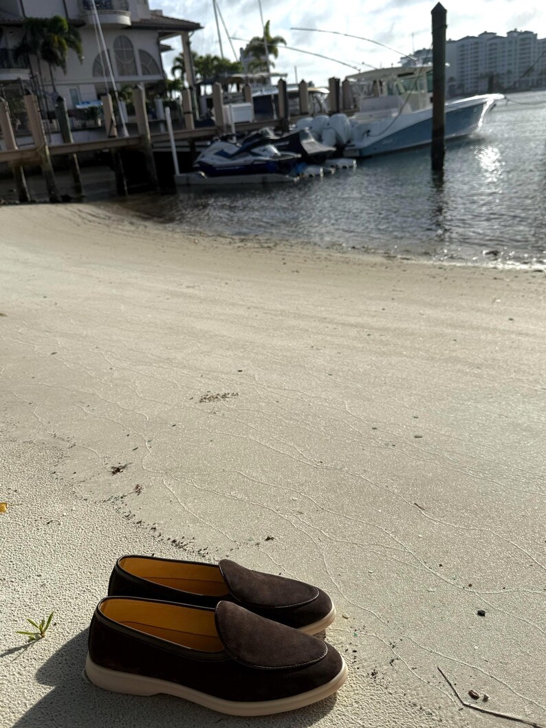 May include: A pair of dark brown suede loafers with a light-colored sole rests on a sandy beach. The shoes have a classic slip-on design, suggesting casual wear. The background shows a waterfront scene with boats and buildings.