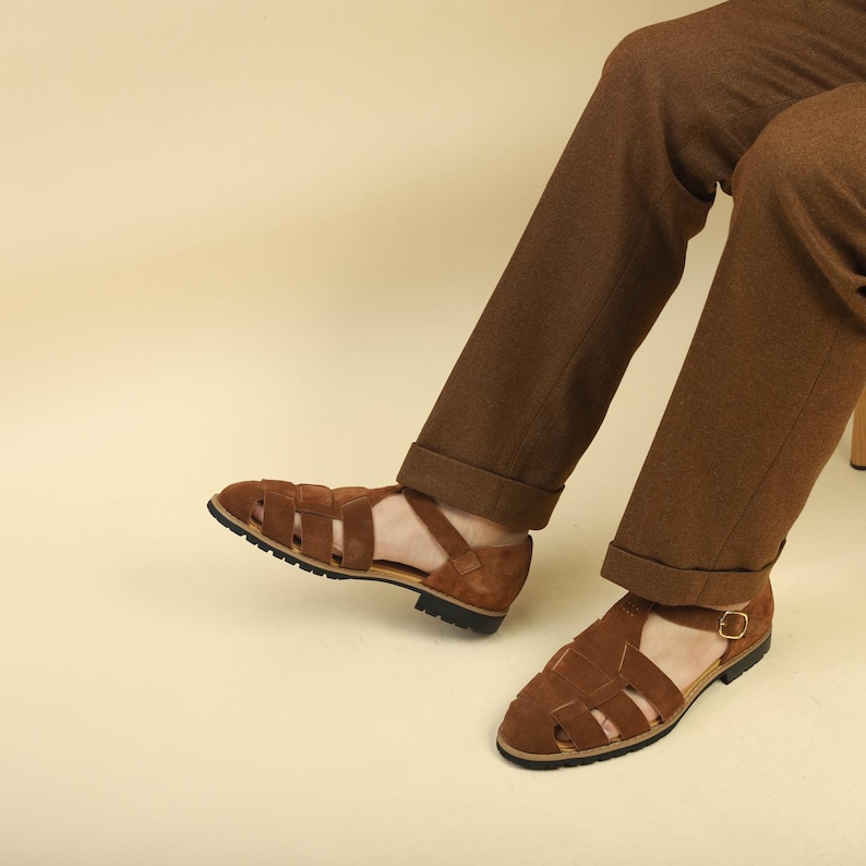 May include: Brown suede sandals with a woven design and a black sole. The sandals are worn with brown pants. The sandals have a strap that buckles on the side.