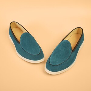Op de afbeelding: Een paar teal su&egrave;de loafers met een ronde neus. De schoenen hebben een zwarte rand en een witte zool. De binnenkant is lichtbruin. De schoenen worden gepresenteerd op een lichtoranje achtergrond. Ongeveer 25 cm lang.