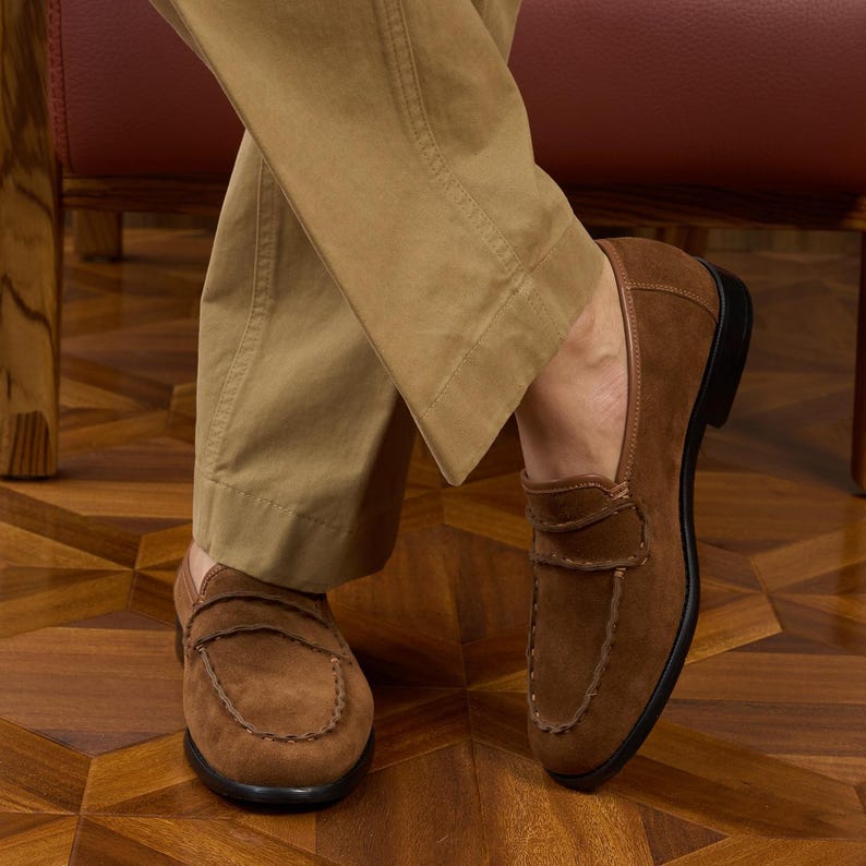 May include: A pair of brown suede penny loafers featuring a classic moccasin-style stitched toe seam, a horizontal strap across the vamp, and a low black heel. The shoes are worn with tan trousers, showcasing the soft texture of the suede material.