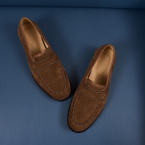 May include: A pair of brown suede loafers featuring a moccasin-style stitched toe seam, a horizontal strap across the vamp, and a smooth tan interior lining. The shoes have a rounded toe shape and a low-profile design with a dark sole edge.