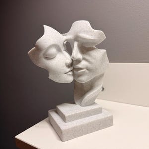 Pode incluir: Escultura branca retratando dois rostos se beijando, um com os olhos fechados. A escultura é montada em uma base escalonada. A obra é feita de um material salpicado semelhante a pedra.