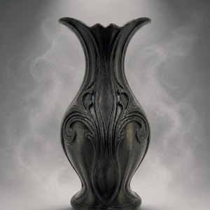 May include: A matte black vase featuring a flared rim and a bulbous body adorned with raised, symmetrical scrollwork patterns that extend from the base toward the neck, creating a textured surface with deep grooves and curved relief details.