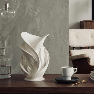 Op de afbeelding: Een witte keramische vaas met een vloeiend, abstract ontwerp dat op bloemblaadjes lijkt. Hij staat op een donker houten oppervlak, naast een kleine witte koffiekop op een zwarte schotel. Een heldere glazen karaf en een gouden lepel zijn ook zichtbaar.