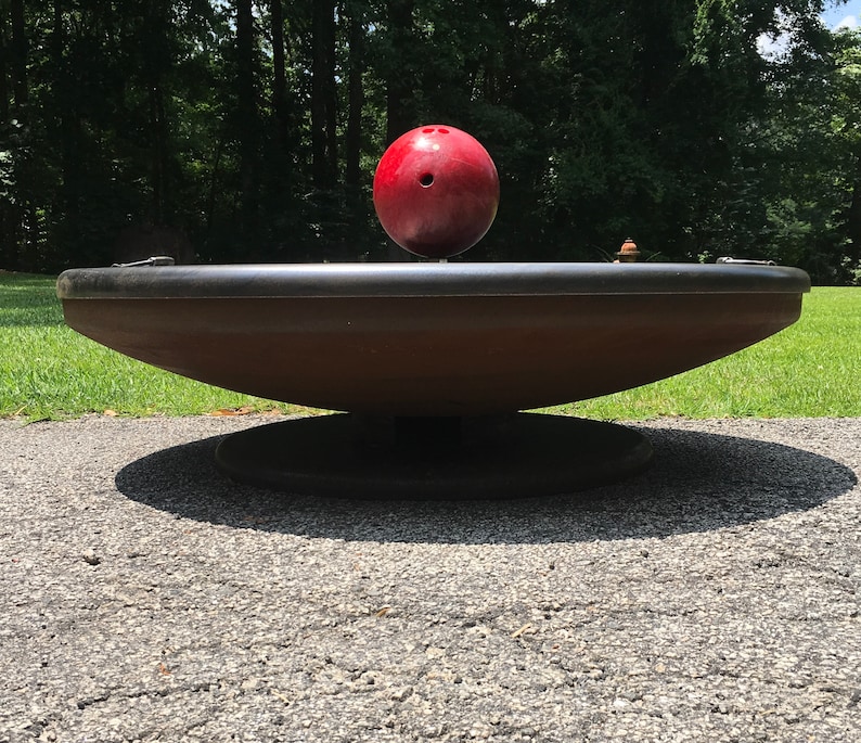 May include: A red ball sits on top of a large, black, circular, spinning platform. The platform is made of metal and has a slightly raised edge. The platform is sitting on a bed of gravel.