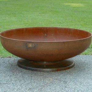 Op de afbeelding: Een grote, roestige metalen vuurkorf met een brede, ronde kom en een verhoogde basis. De vuurkorf staat op een grijs, geplaveid oppervlak.