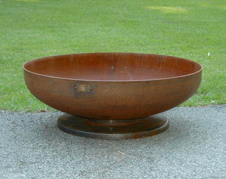 Op de afbeelding: Een grote, roestige metalen vuurkorf met een brede, ronde kom en een verhoogde basis. De vuurkorf staat op een grijs, geplaveid oppervlak.