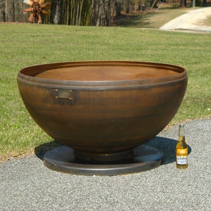 May include: A large, round, rusty-brown metal fire pit with a wide bowl shape, set on a dark circular base. A small rectangular metal plate is attached to the side. A bottle of beer sits next to the fire pit.