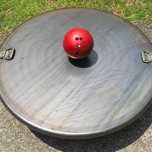 May include: A red bowling ball sits on a large, round, metal disc with two metal handles. The disc has a textured surface.